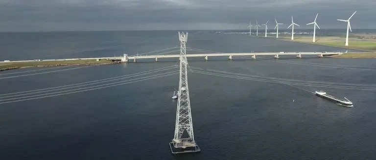 Hoogspanningsmast in het water bij Flevoland met op de achtergrond een brug en een rij windturbines.